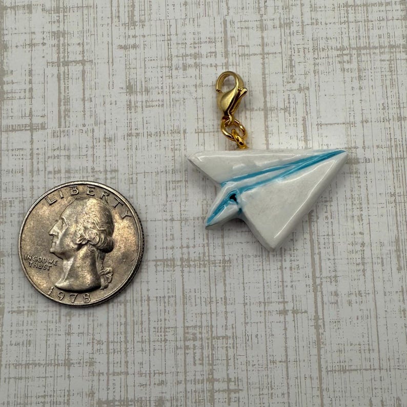 May include: A white paper airplane charm with blue accents and a gold-colored clasp. The charm is next to a quarter coin. The charm is likely for a bracelet or necklace.