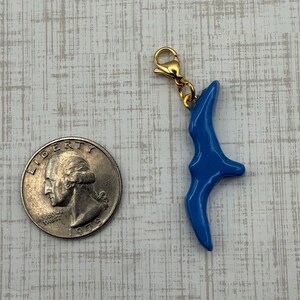 May include: A blue charm with a gold-colored clasp. The charm has a curved, abstract shape. A quarter coin is next to the charm for size comparison.