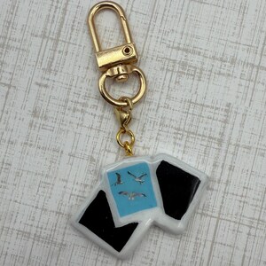 May include: A gold-colored keychain with a rectangular charm. The charm features a light blue square with three seagulls and two black squares. The keychain has a gold clasp.