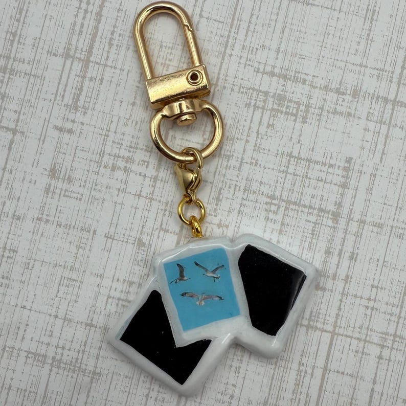 May include: A gold-colored keychain with a rectangular charm. The charm features a light blue square with three seagulls and two black squares. The keychain has a gold clasp.