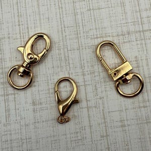 May include: Three gold-colored swivel clasps of varying sizes. The clasps have a lobster claw design with a spring-loaded gate and a circular ring for attaching to other items. The clasps are on a light-colored, textured surface.