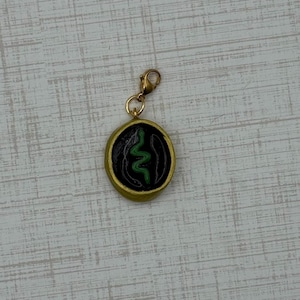 May include: A gold-colored charm with a black center featuring a green snake design. The charm is round with a gold border and a gold clasp. The snake design is a stylized, wavy line.