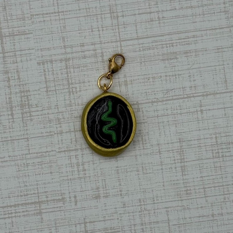 May include: A gold-colored charm with a black center featuring a green snake design. The charm is round with a gold border and a gold clasp. The snake design is a stylized, wavy line.