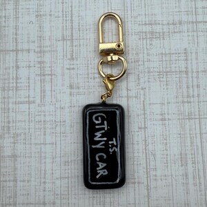 May include: A black rectangular keychain with the text "GTWY CAR" and "T.S." in white. The keychain has a gold-colored clasp and a small gold-colored ring.