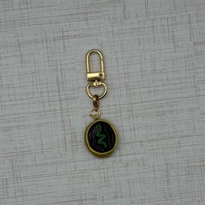 May include: Gold-colored keychain with a round pendant featuring a green snake design on a black background. The pendant has a gold border. The keychain has a clasp for easy attachment.