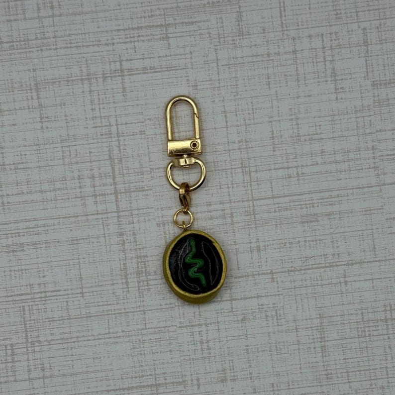 May include: Gold-colored keychain with a round pendant featuring a green snake design on a black background. The pendant has a gold border. The keychain has a clasp for easy attachment.
