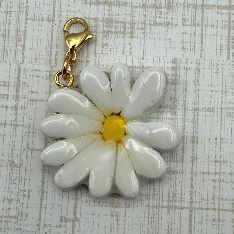Op de afbeelding: Een witte madeliefjesbedel met een gele kern en een goudkleurige sluiting. Het madeliefje heeft meerdere witte bloemblaadjes en een glanzende afwerking. De bedel is ontworpen om aan een armband of ketting te worden bevestigd.