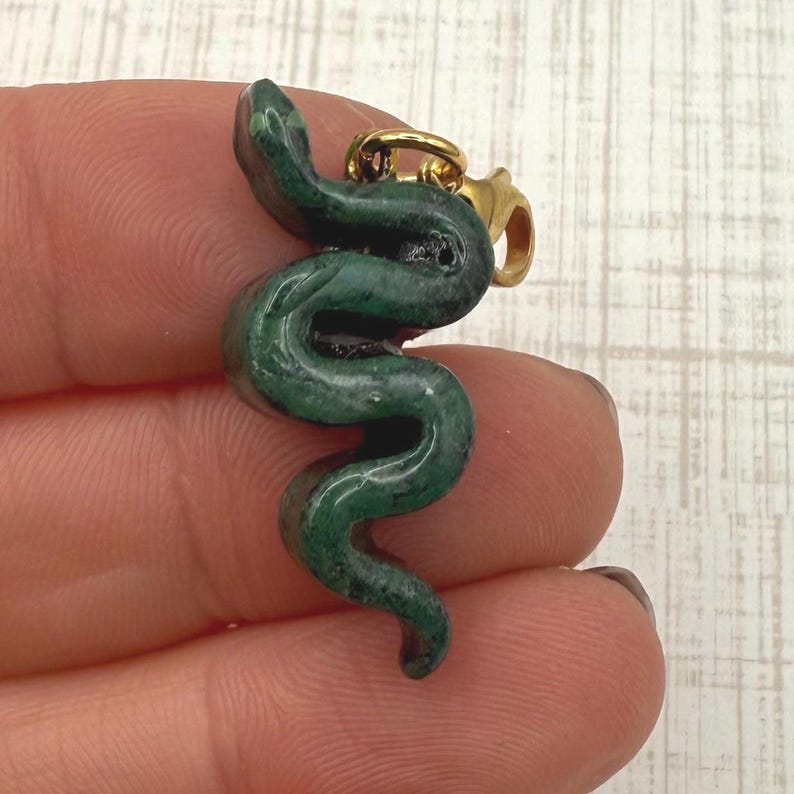 May include: A green serpentine pendant in the shape of a snake. The snake is coiled and has a gold-colored clasp. The pendant is held between two fingers.