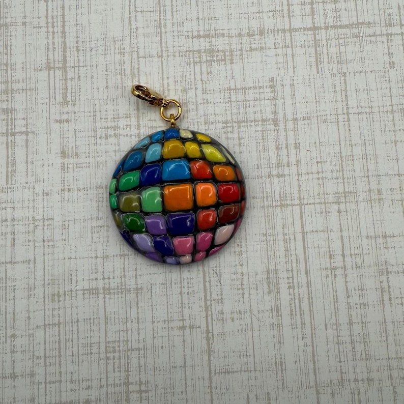 Puede incluir: Un dije redondo y colorido con un cierre dorado. El dije es un mosaico de colores vibrantes de forma cuadrada, incluyendo azul, verde, naranja, rojo y morado. El dije es probablemente para un collar o pulsera.