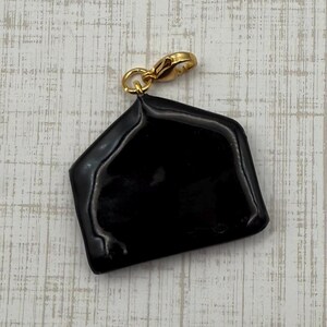 May include: A black, glossy charm with a gold-colored clasp. The charm is a geometric shape, possibly a stylized house or envelope. The clasp is attached to the top of the charm, allowing it to be added to a necklace or bracelet.