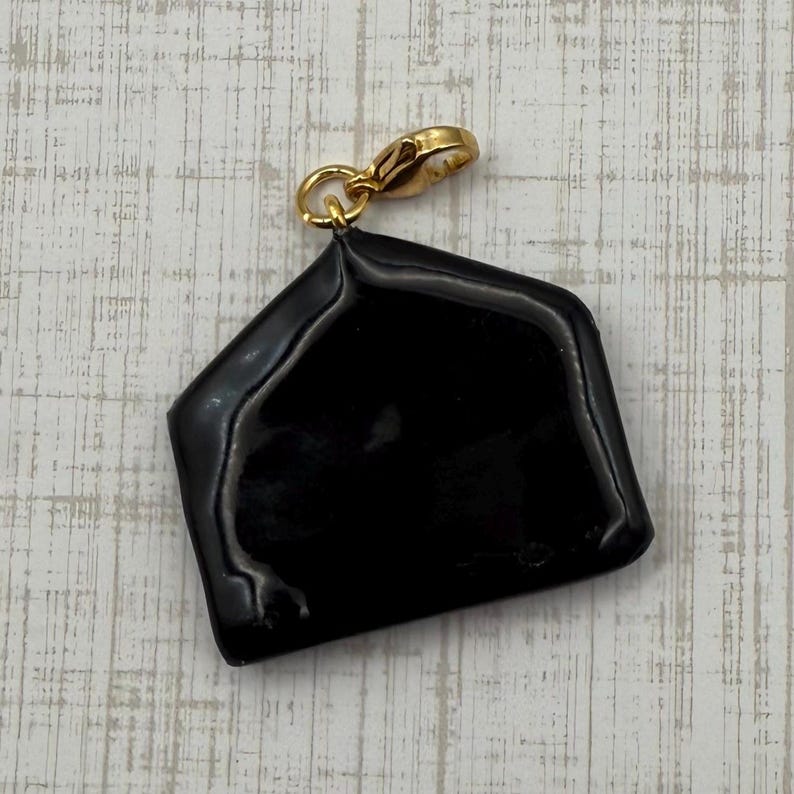 May include: A black, glossy charm with a gold-colored clasp. The charm is a geometric shape, possibly a stylized house or envelope. The clasp is attached to the top of the charm, allowing it to be added to a necklace or bracelet.