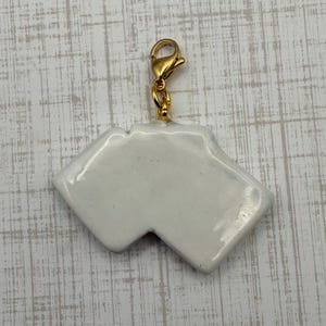 May include: A white ceramic charm with a gold-colored clasp. The charm has a unique shape with two angled sections and a notch. The charm is likely for a bracelet or necklace.