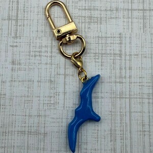 May include: A gold-colored keychain with a blue bird-shaped charm. The keychain has a lobster clasp and a split ring. The bird charm is a vibrant blue color and has a smooth, glossy finish. The background is a light gray and white wood grain pattern.