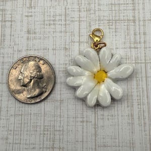 Op de afbeelding: Een witte madeliefjesbedel met een gele kern en een goudkleurige sluiting. De bedel staat naast een Amerikaanse kwartmunt ter vergelijking van de grootte. De madelief heeft witte bloemblaadjes en een gele kern.