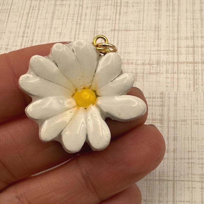 Op de afbeelding: Een witte madeliefhanger met een gele kern en een goudkleurige beugel. De madelief heeft meerdere witte bloemblaadjes en wordt in een hand gehouden. De hanger is een accessoire met een bloemig thema.