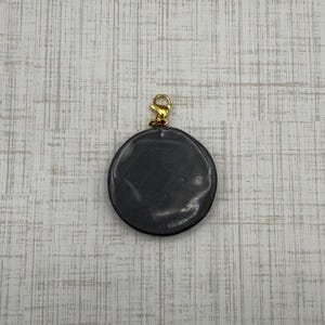 Puede incluir: Un colgante redondo gris oscuro con un cierre dorado. El colgante tiene una superficie lisa y brillante y est&aacute; suspendido de un peque&ntilde;o cierre de mosquet&oacute;n dorado. El fondo es un patr&oacute;n de vetas de madera gris claro y blanco.