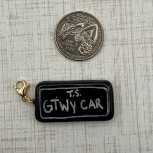 May include: A black rectangular charm with the text "T.S. GTWY CAR" in white lettering. The charm has a gold-colored clasp. A quarter is next to the charm.