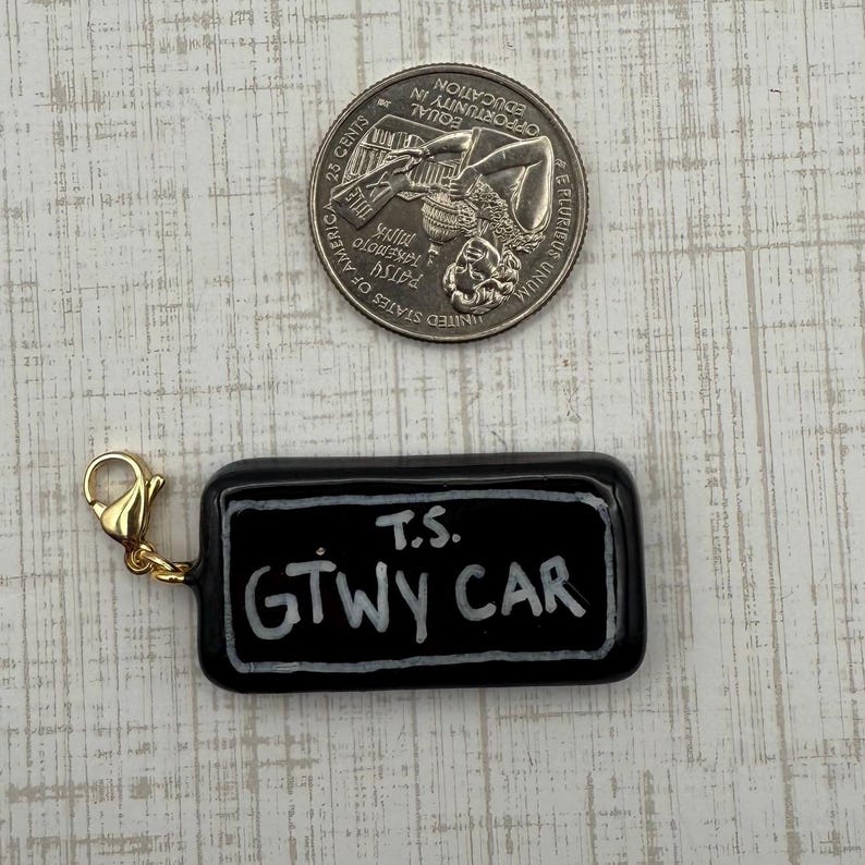 May include: A black rectangular charm with the text "T.S. GTWY CAR" in white lettering. The charm has a gold-colored clasp. A quarter is next to the charm.