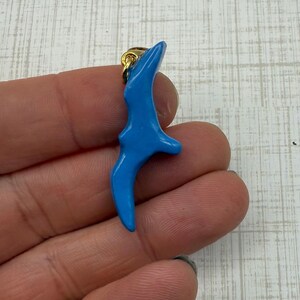 May include: A bright blue pendant in the shape of a bird in flight, with a gold-colored bail. The pendant is smooth and glossy, and the bird's form is stylized. The pendant is held in a hand.