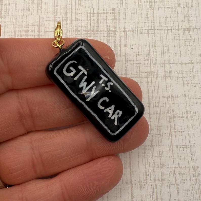 May include: A black rectangular charm with the text "GTWy CAR" in white lettering. The charm has a gold-colored clasp at the top. The charm is held in a person's hand.