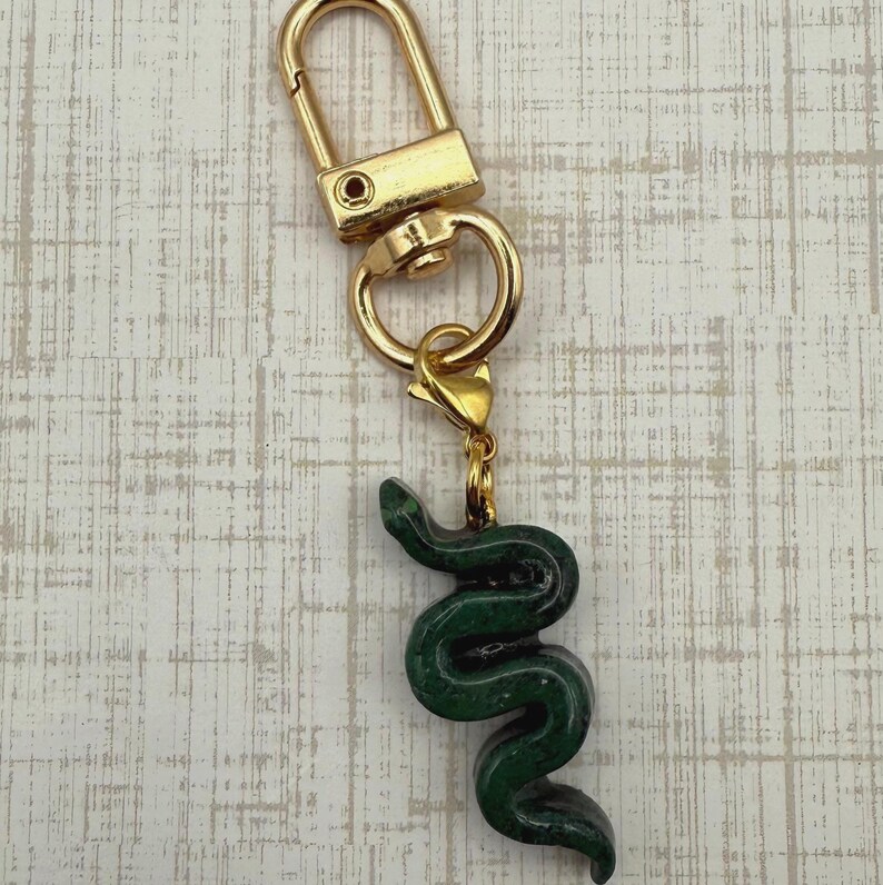 May include: A gold-colored keychain with a green snake charm. The snake charm is detailed and has a textured appearance. The keychain has a clasp for easy attachment to bags or keys.