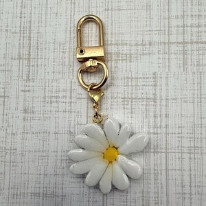 Op de afbeelding: Een goudkleurige sleutelhanger met een witte madeliefjesbedel. Het madeliefje heeft een gele kern en witte bloemblaadjes. De sleutelhanger heeft een karabijnhaak voor eenvoudige bevestiging.