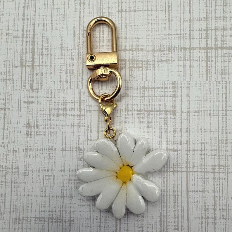 Op de afbeelding: Een goudkleurige sleutelhanger met een witte madeliefjesbedel. Het madeliefje heeft een gele kern en witte bloemblaadjes. De sleutelhanger heeft een karabijnhaak voor eenvoudige bevestiging.