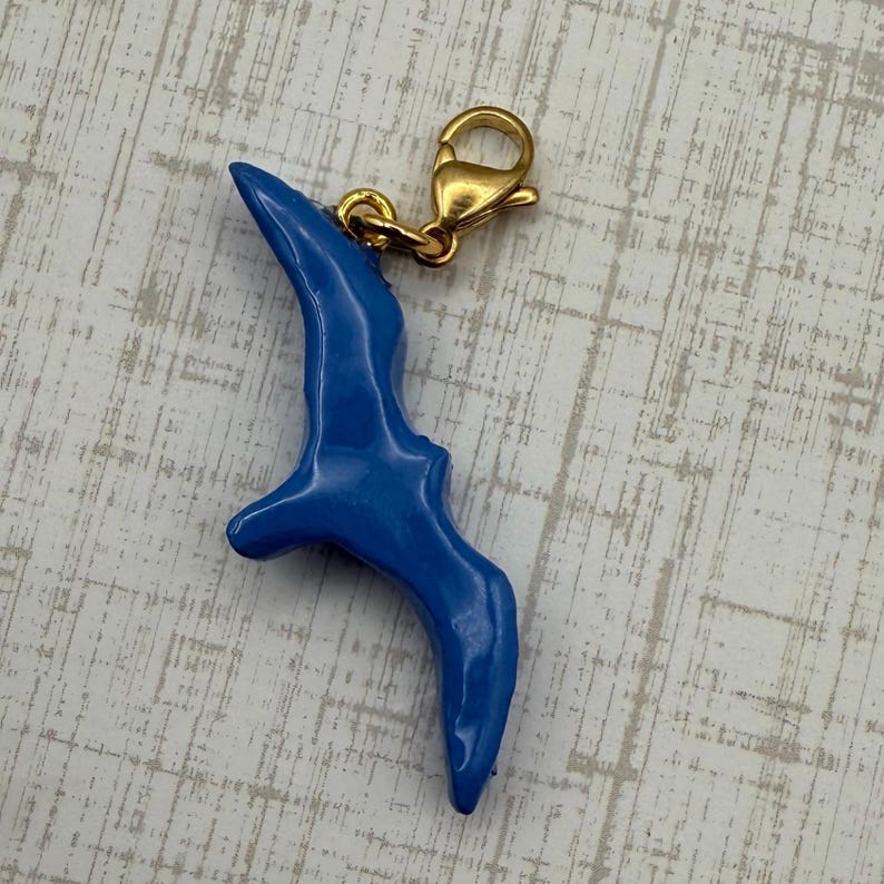 May include: A glossy, blue bird charm with a gold-colored clasp. The bird charm is a stylized representation of a bird in flight. The charm is attached to a gold-colored lobster clasp.