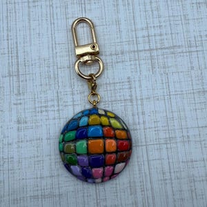Puede incluir: Un llavero redondo y colorido con un cierre dorado. El dije es un mosaico de baldosas cuadradas en varios colores, incluyendo azul, verde, naranja y morado. El llavero est&aacute; sujeto a un cierre met&aacute;lico dorado.