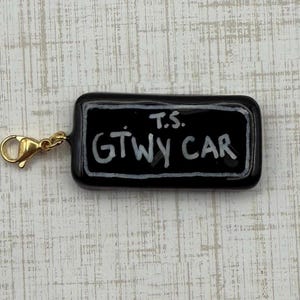 May include: A black rectangular charm with the text "T.S. GTWY CAR" in white, attached to a gold-colored clasp. The charm has a glossy finish and a white border around the text.