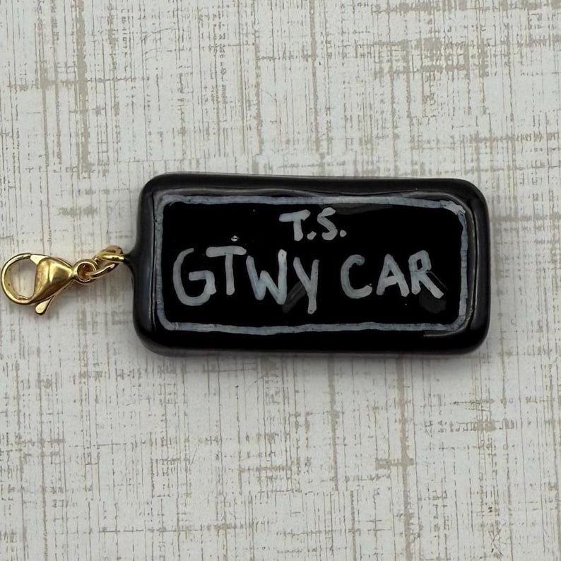 May include: A black rectangular charm with the text "T.S. GTWY CAR" in white, attached to a gold-colored clasp. The charm has a glossy finish and a white border around the text.