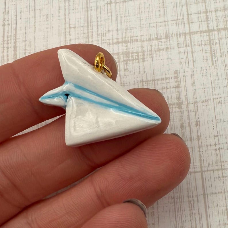 May include: A white ceramic paper airplane pendant with blue stripes and a gold-colored loop for hanging. The pendant is held in a hand, showcasing its delicate design and craftsmanship. This unique jewelry piece is perfect for aviation enthusiasts.