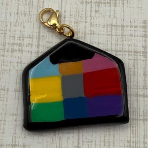 May include: A colorful house-shaped charm with a gold-colored clasp. The charm features a black outline and a patchwork design of various colors, including blue, yellow, green, red, and purple. The charm is likely made of resin or a similar material.
