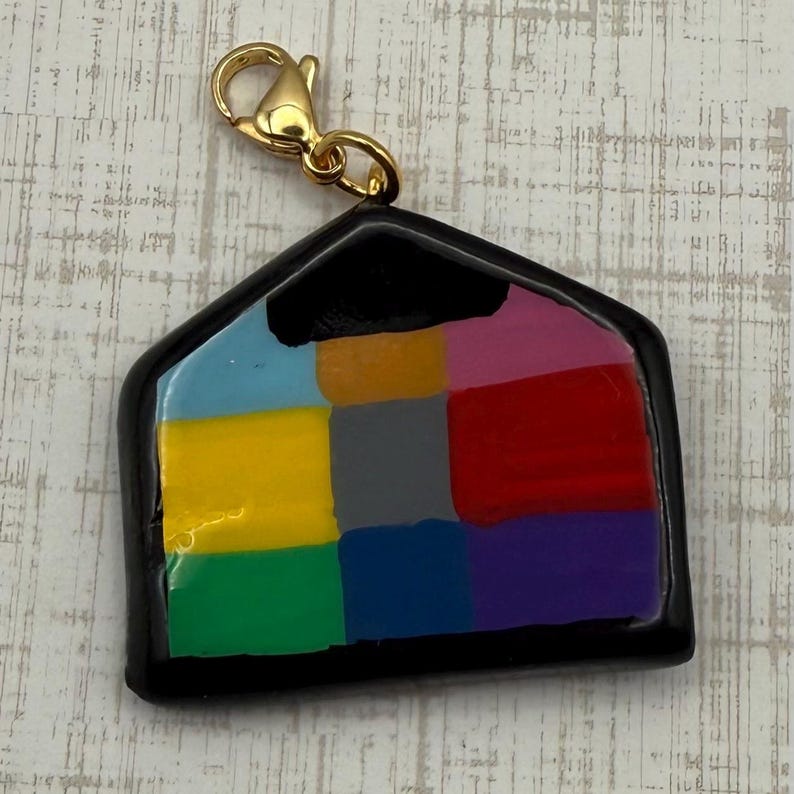 May include: A colorful house-shaped charm with a gold-colored clasp. The charm features a black outline and a patchwork design of various colors, including blue, yellow, green, red, and purple. The charm is likely made of resin or a similar material.