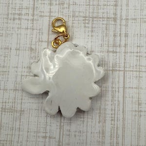 Op de afbeelding: Een witte bloemvormige bedel met een goudkleurige sluiting. De bedel heeft een glanzende afwerking en een gedetailleerd, bloembladachtig ontwerp. De sluiting is bevestigd aan de bovenkant van de bedel.
