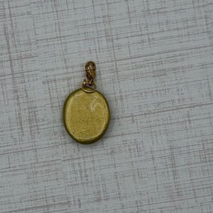 May include: A gold-colored oval charm with a small clasp. The charm has a textured surface and a simple design. The clasp is also gold-colored and appears to be made of metal. The charm is likely for a necklace or bracelet.