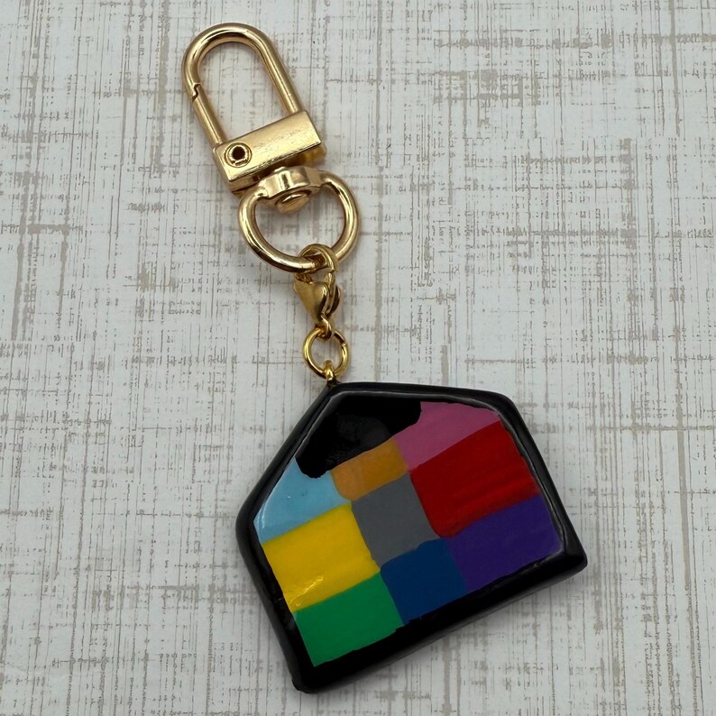 May include: A colorful keychain with a gold-tone clasp. The pendant is a black, geometric shape filled with squares of various colors, including blue, yellow, red, and purple. The keychain is likely made of resin or a similar material.