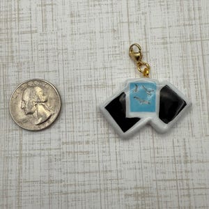 May include: A white and black charm with a gold clasp. The charm features a light blue square with three seagulls and two black squares. A U.S. quarter is next to the charm.