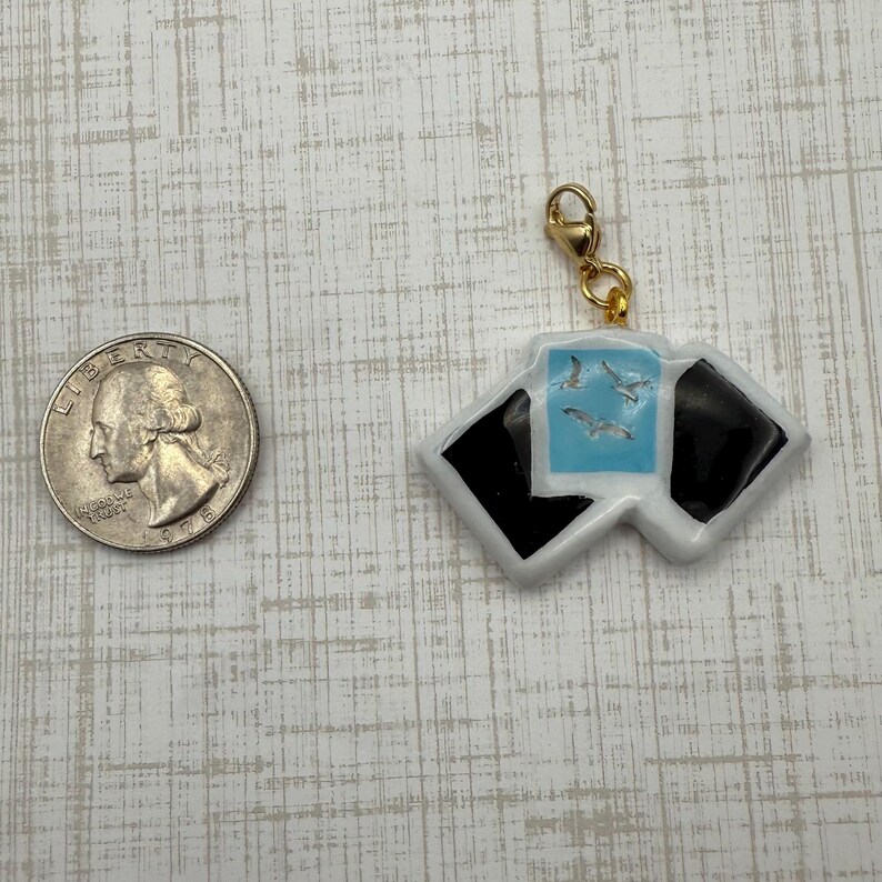 May include: A white and black charm with a gold clasp. The charm features a light blue square with three seagulls and two black squares. A U.S. quarter is next to the charm.