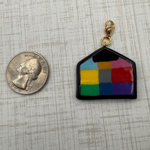 May include: A colorful charm in the shape of a house with a gold-colored clasp. The charm is divided into nine squares of different colors: blue, yellow, green, red, purple, pink, and gray. A U.S. quarter is next to the charm.
