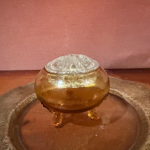 May include: A round, amber-colored glass bowl featuring three small feet and a clear glass flower frog insert with multiple circular openings at the top. The bowl has a smooth, curved body and a translucent finish, designed for holding floral arrangements.