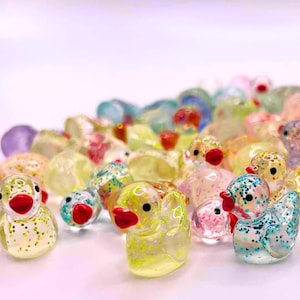 May include: A selection of colourful glass duck figurines with red beaks and black eyes. The ducks are in various colours, including yellow, blue, green, and pink, with glitter accents. The figurines are scattered on a white surface.
