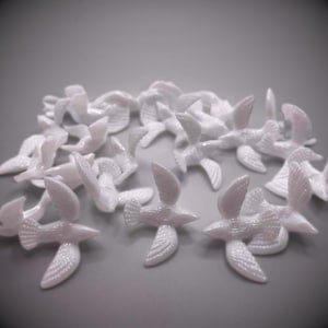 May include: A group of small, white, bird-shaped ornaments. Each bird has outstretched wings and a detailed tail, suggesting a flock in flight. The objects are arranged on a grey surface.