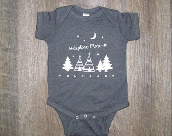 May include: A dark gray baby onesie with a white graphic of a crescent moon, stars, trees, teepees, and the text "Explore More - Colorado".