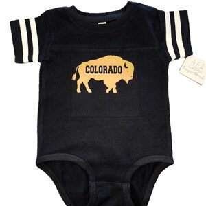 May include: Black baby onesie with white stripes on the sleeves. The onesie has a gold silhouette of a buffalo with the word "COLORADO" printed on it.