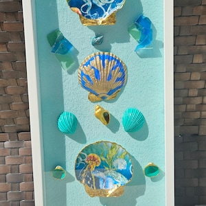May include: A framed art piece featuring seashells and sea-themed elements. The artwork includes painted seashells with gold accents, sea glass, and a starfish, all arranged on a light blue background within a white frame.