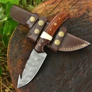 May include: A handcrafted hunting knife with a dark wood handle, brass rivets, and a bone spacer. The blade is made of patterned steel and has a curved tip. It comes with a brown leather sheath and belt loops with brass buttons.