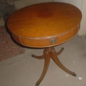 May include: Oval wooden side table with a warm, medium-brown finish. The table features a single drawer with a brass pull and is supported by a tripod base with curved legs. The table's dimensions are approximately 24 inches in diameter and 28 inches high.