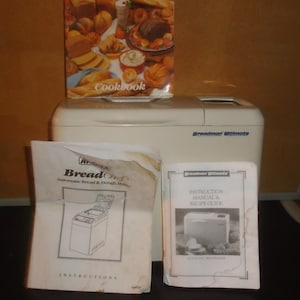 May include: A white Breadman Ultimate bread maker with a cookbook and instruction manuals. The cookbook features images of various breads. The instruction manuals are for the Bread Chef Automatic Bread & Dough Maker.