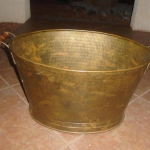 May include: A hammered brass bucket with wooden handles. The bucket has a wide, oval shape and a textured surface. The handles are attached to the sides with metal brackets. The bucket is a warm, golden color.